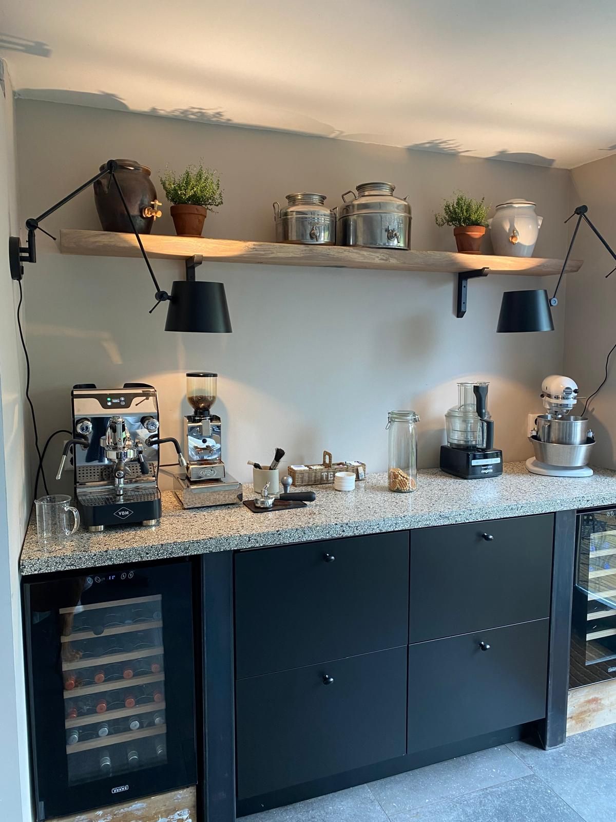 De coffee corner: espressomachine en koffiemolen op een granieten blad boven zwarte kasten, met een eiken plank vol olijfoliekannen en tijm