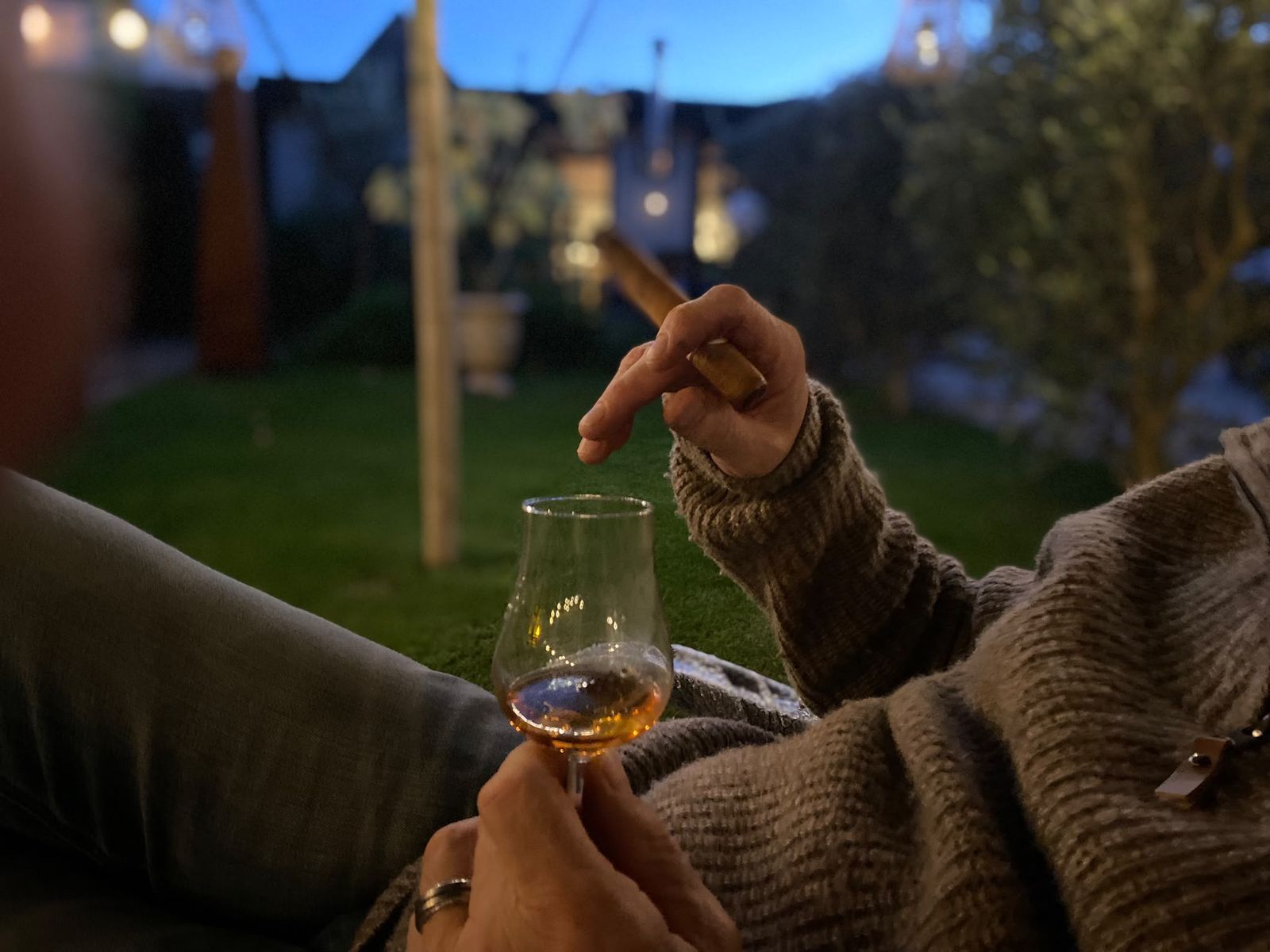 Een glas whisky in de tuin bij schemer, met de lampjes van de stretchtent op de achtergrond